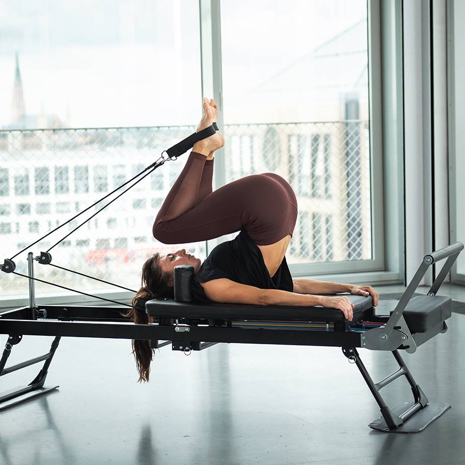 Kobieta ćwiczy Pilates na reformerze z nogami uniesionymi i stopami w paskach.