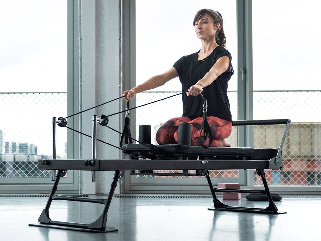 Kobieta ćwiczy Pilates na reformerze, w tle duże okna.