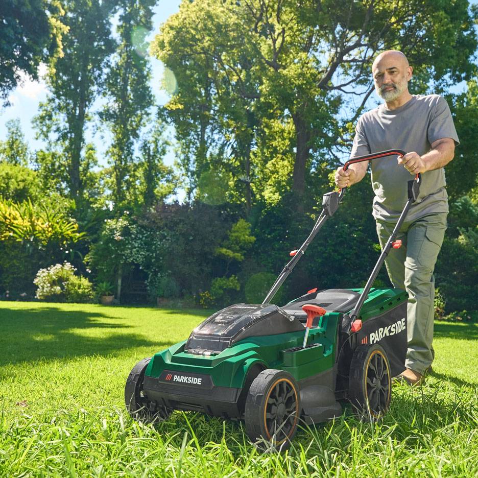 Mężczyzna kosi trawę zielonym wertykulatorem w słonecznym ogrodzie.