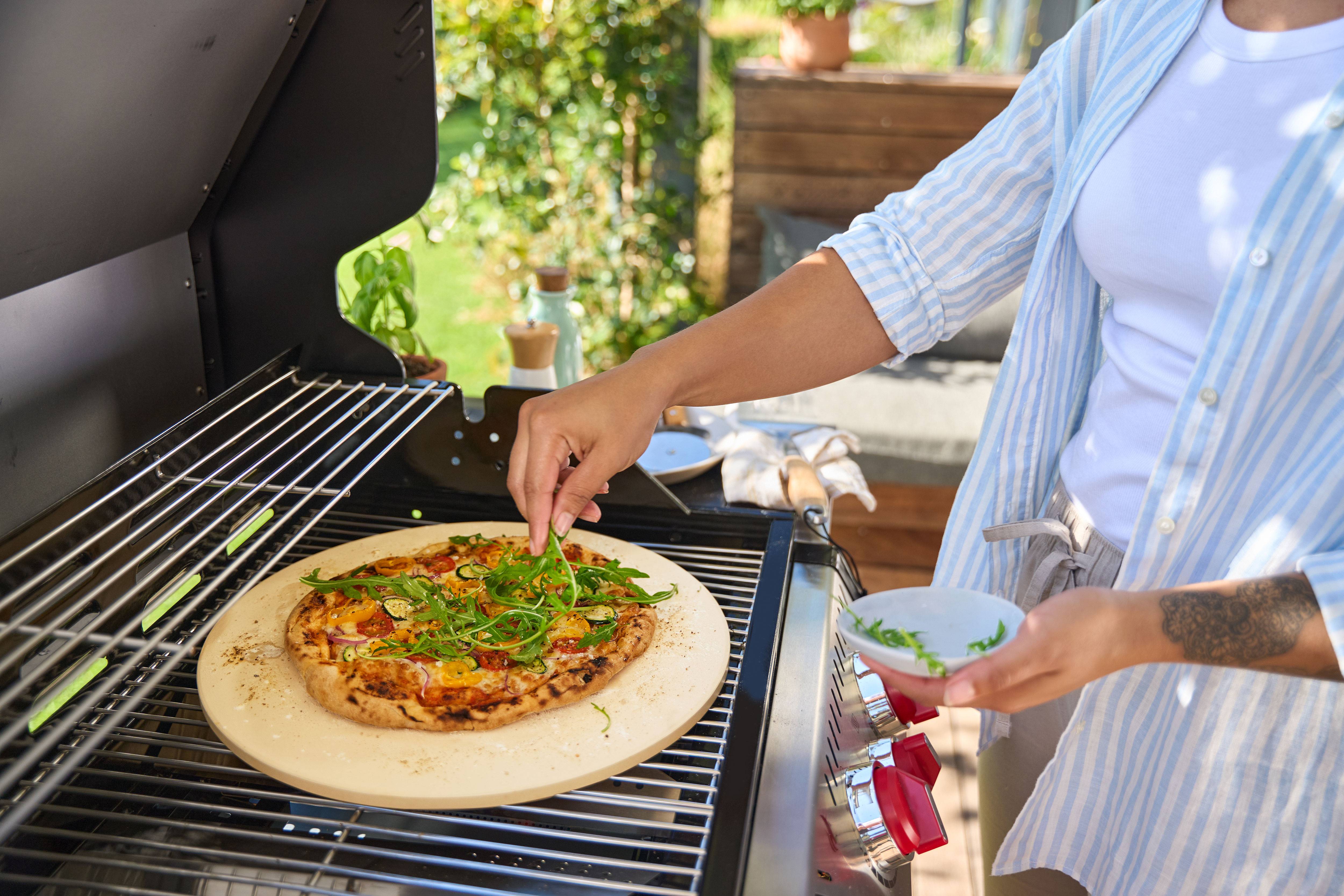 Osoba dodająca rukole do pizzy na kamieniu w grillu gazowym.