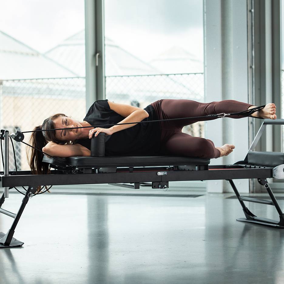 Kobieta ćwiczy Pilates na maszynie reformer, leżąc na boku z paskami na nogach.