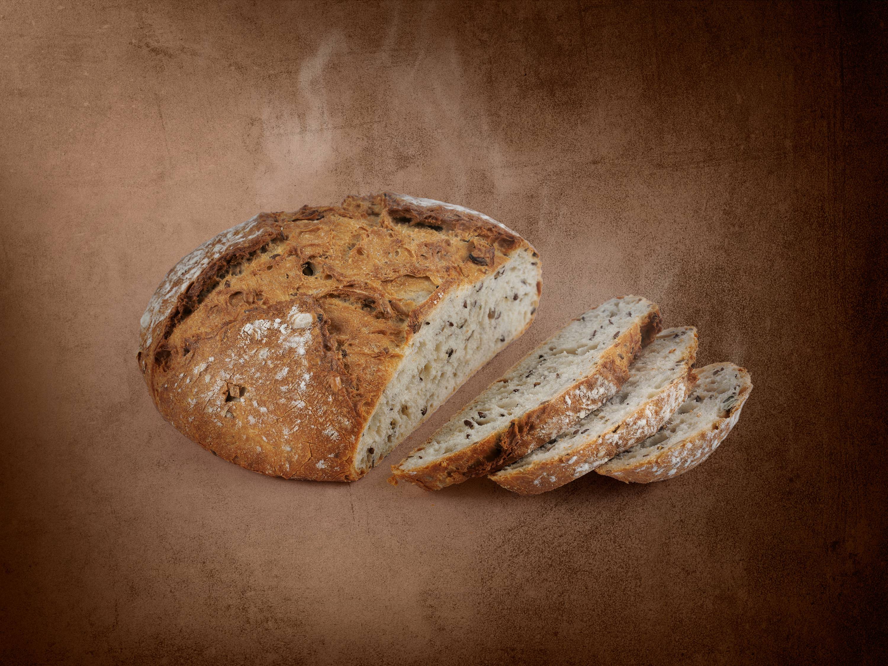 Bochenek świeżego chleba z ziarnami, częściowo pokrojony, na brązowym tle.