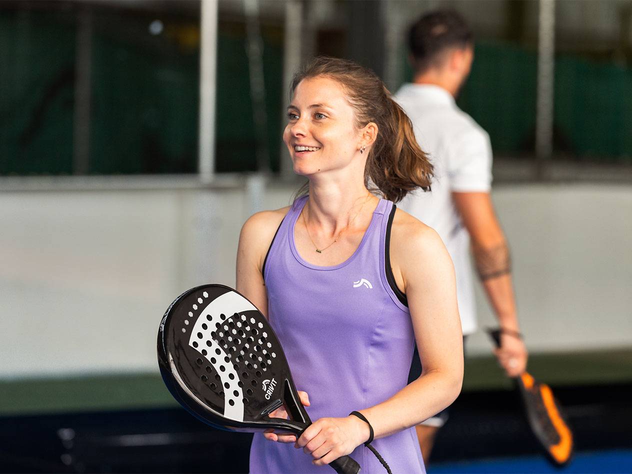 Kobieta w fioletowej sukience sportowej trzyma rakietę do padla, mężczyzna w tle.