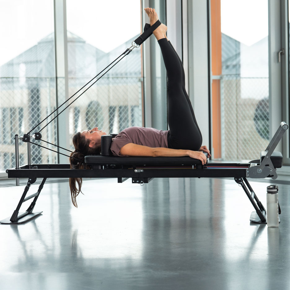 Kobieta ćwiczy Pilates na reformerze z nogami w górze, w jasnym pokoju.