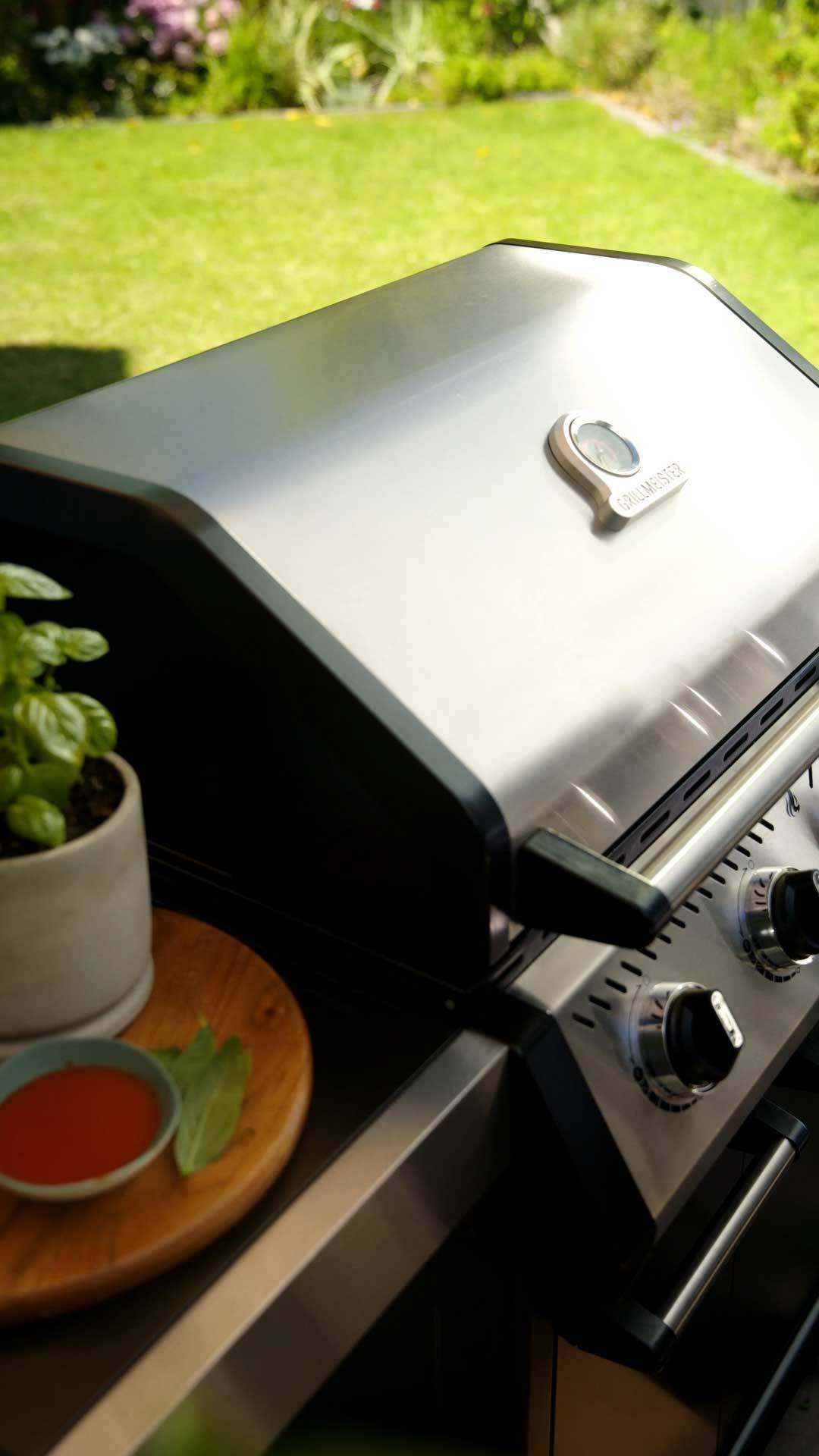 Grill gazowy ze stali nierdzewnej z termometrem, obok doniczki z bazylią i sosem.
