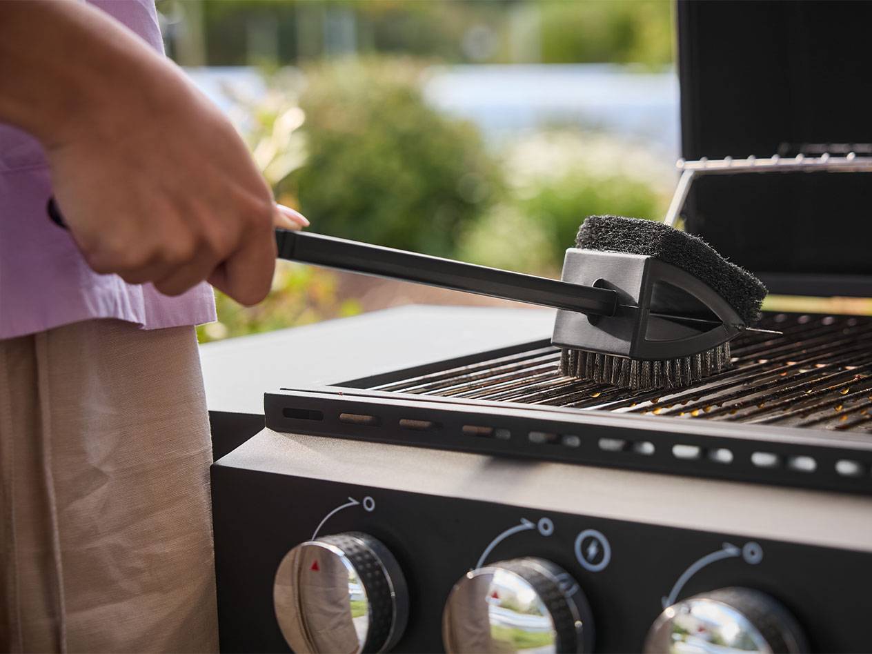 Osoba czyści grill szczotką z długą rączką.