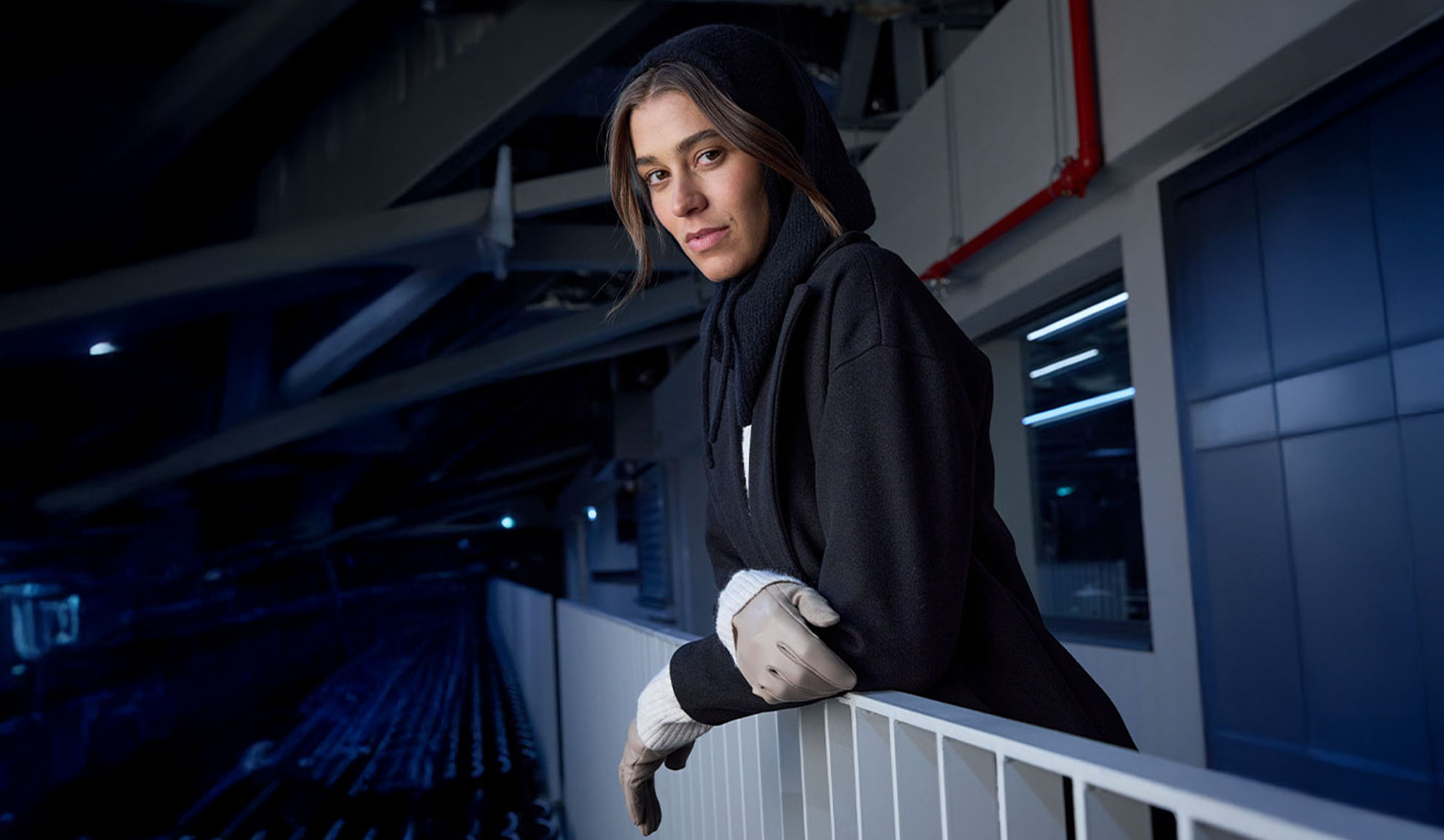 Kobieta w czarnym płaszczu i szaliku z kapturem, opierająca się o barierkę w nowoczesnym otoczeniu.