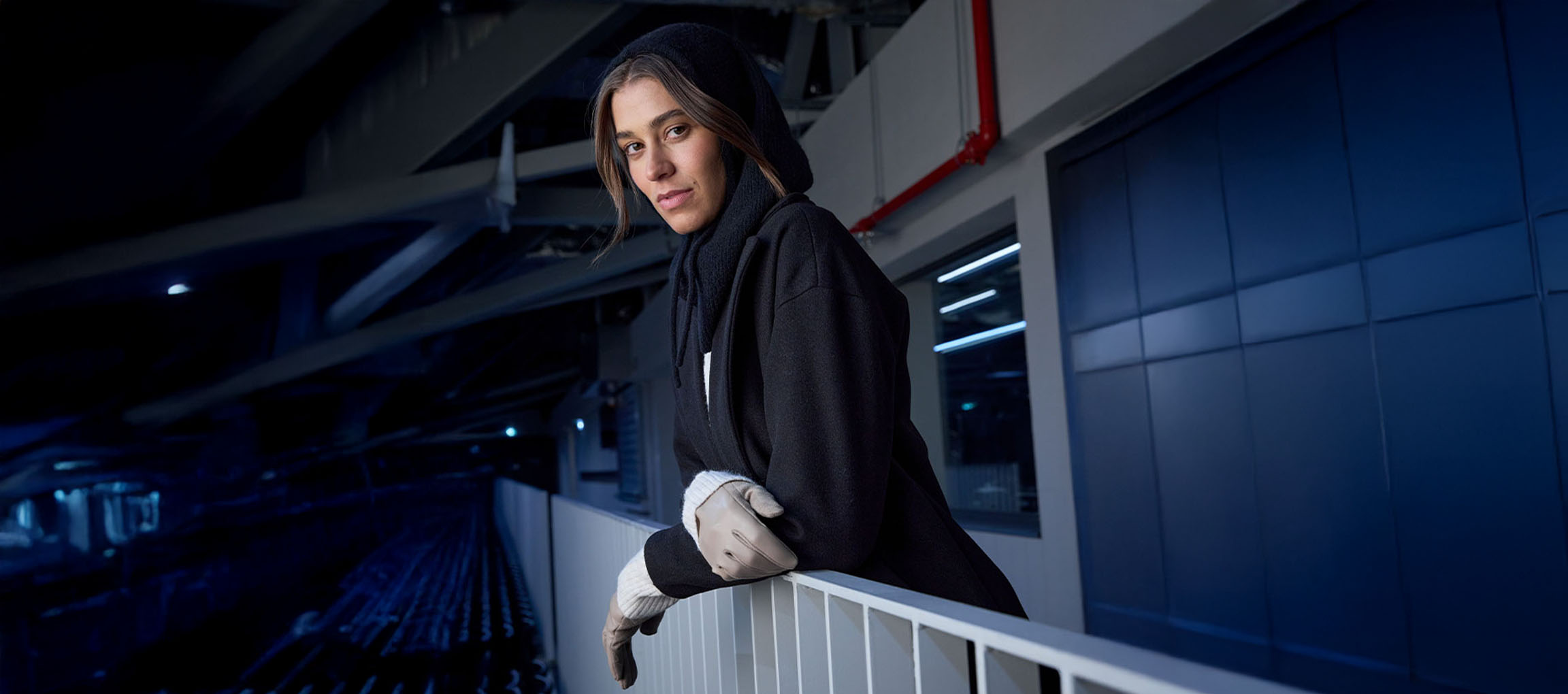 Kobieta w czarnym płaszczu i rękawiczkach oparta o barierkę w miejskim otoczeniu.