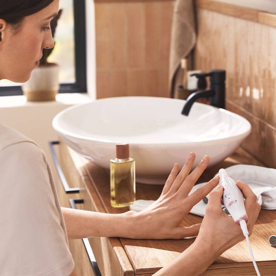 Kobieta używająca elektrycznego zestawu do manicure/pedicure przy drewnianej toaletce z białą umywalką.