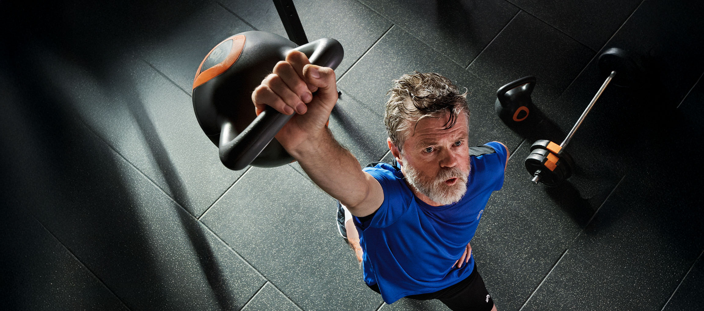 Mężczyzna podnosi kettlebell na siłowni, ze sztangą i kettlebell na podłodze.