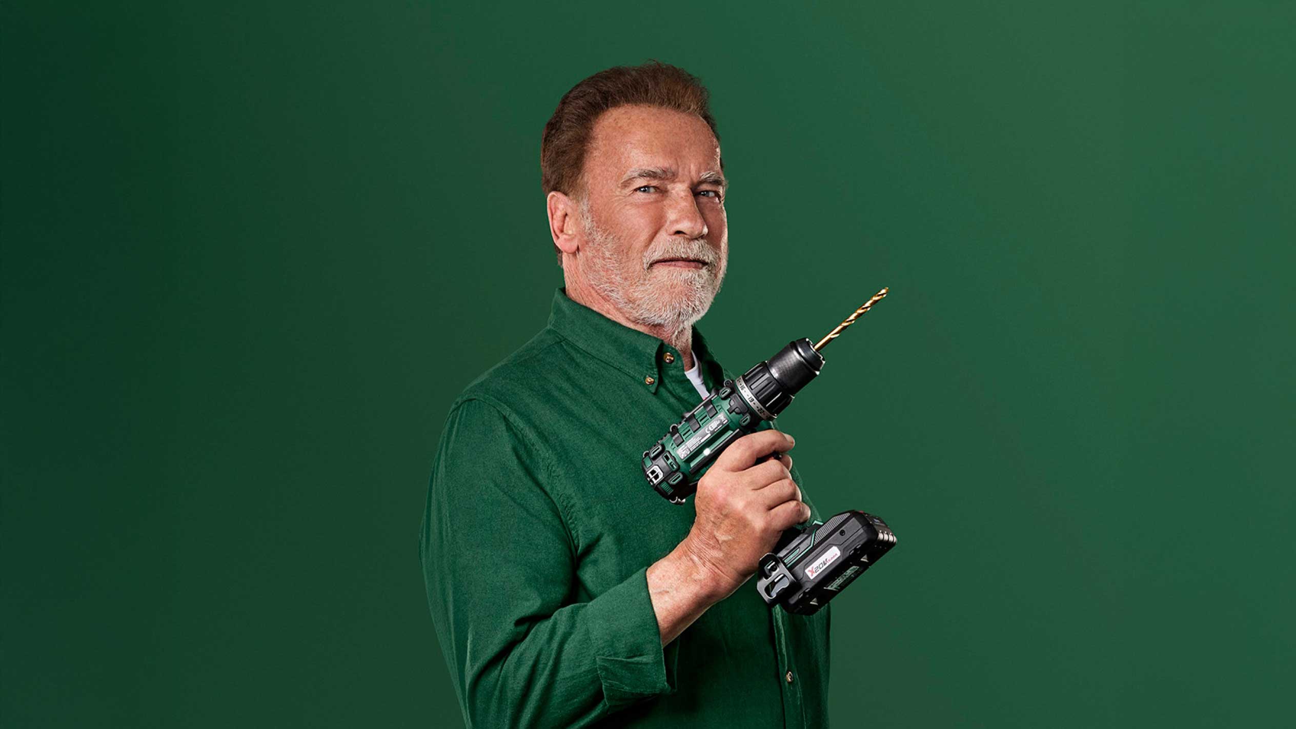 Arnold Schwarzenegger trzyma zieloną wiertarkę akumulatorową ze złotym wiertłem.