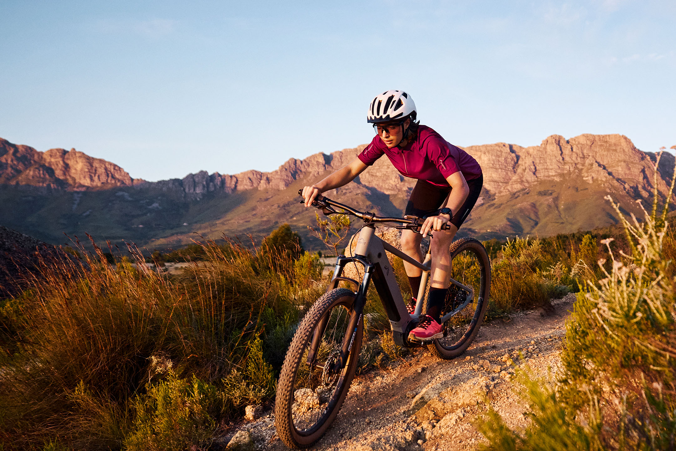 Kobieta na rowerze górskim na szutrowej ścieżce z górami w tle.