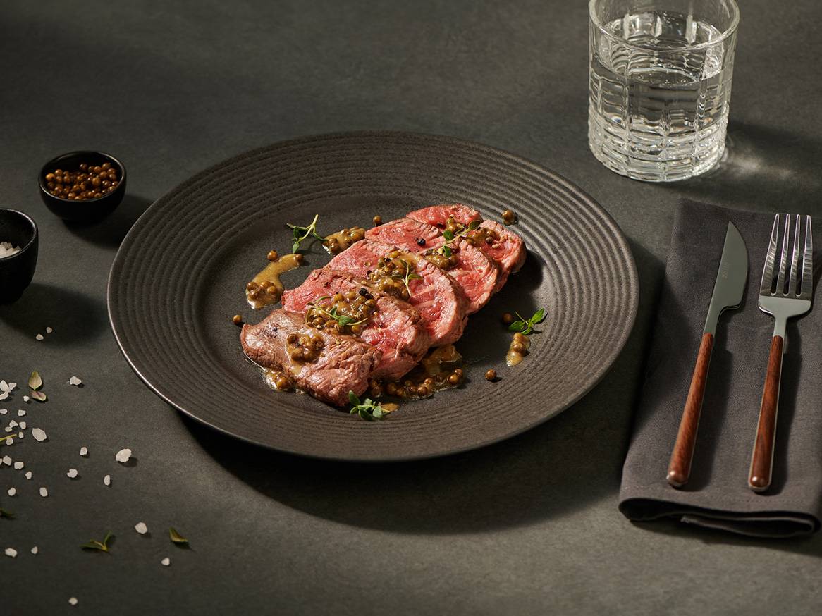 Plasterki pieczonej wołowiny z sosem pieprzowym, podane na ciemnym talerzu z wodą i sztućcami.