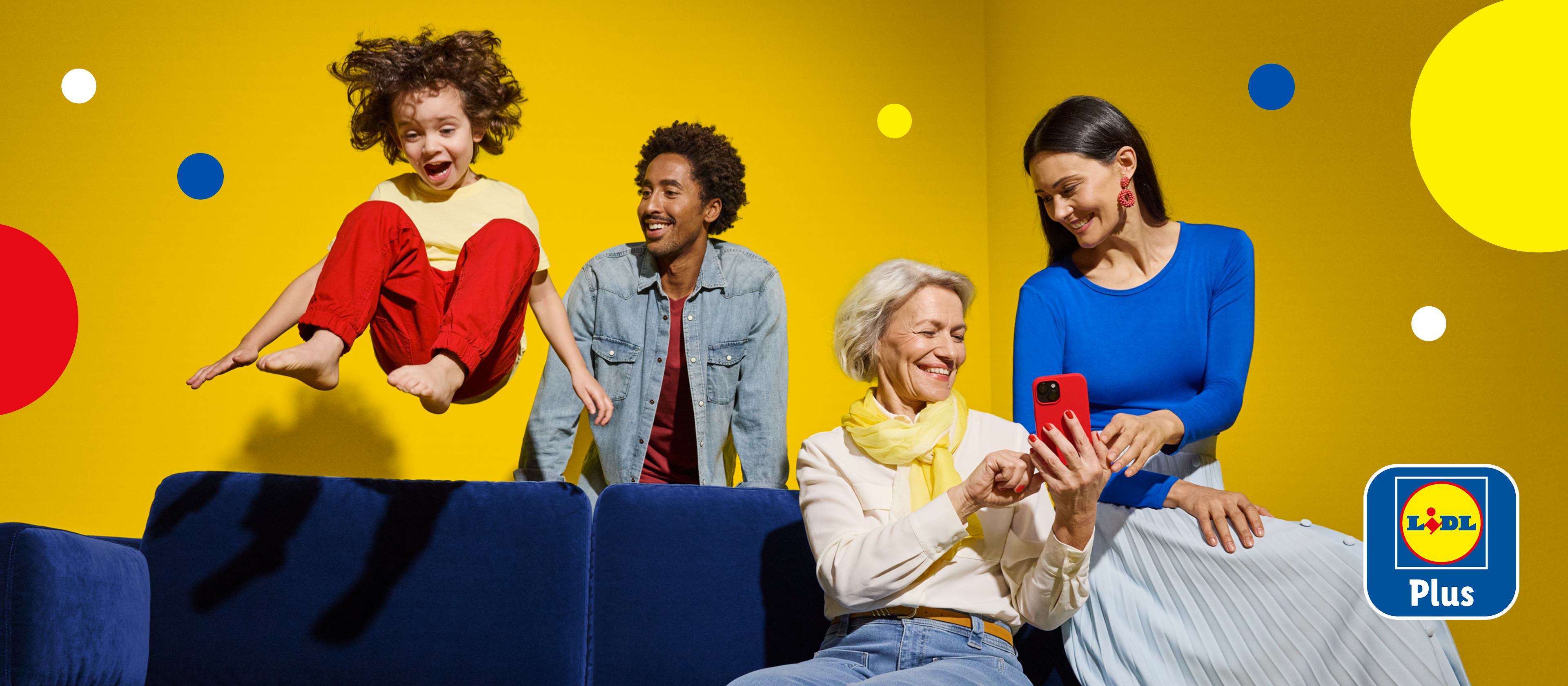 Rodzina na kanapie, dziecko skacze, a dorośli uśmiechają się, patrząc na telefon.