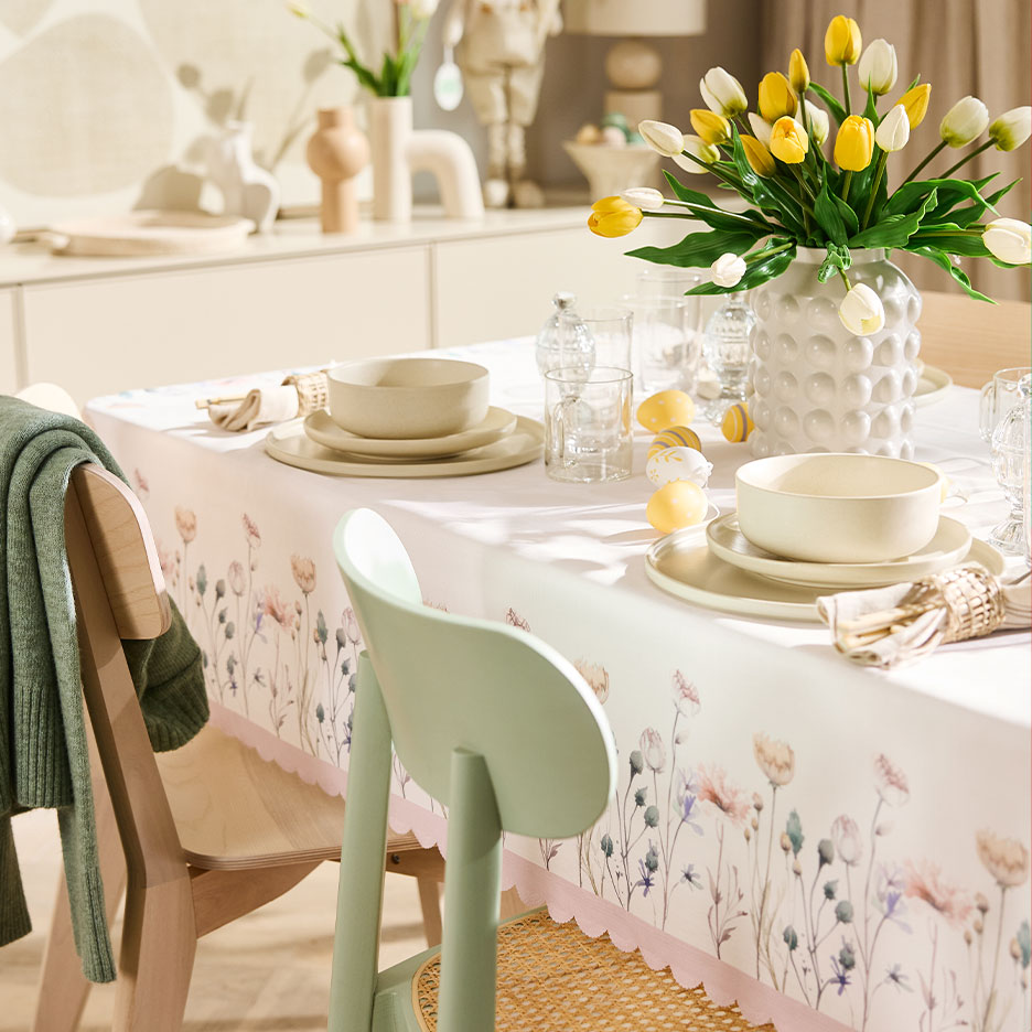 Stół wielkanocny nakryty obrusem w kwiaty, ceramiczną zastawą i żółto-białymi tulipanami.