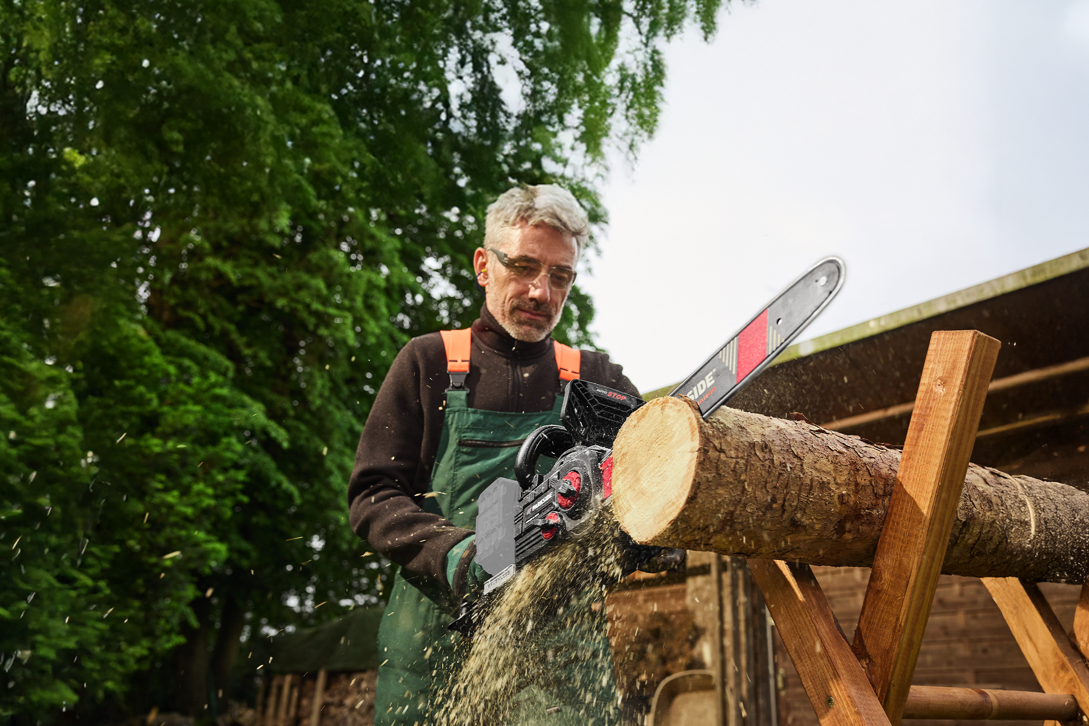 Mężczyzna w okularach ochronnych tnie drewno piłą łańcuchową Parkside.