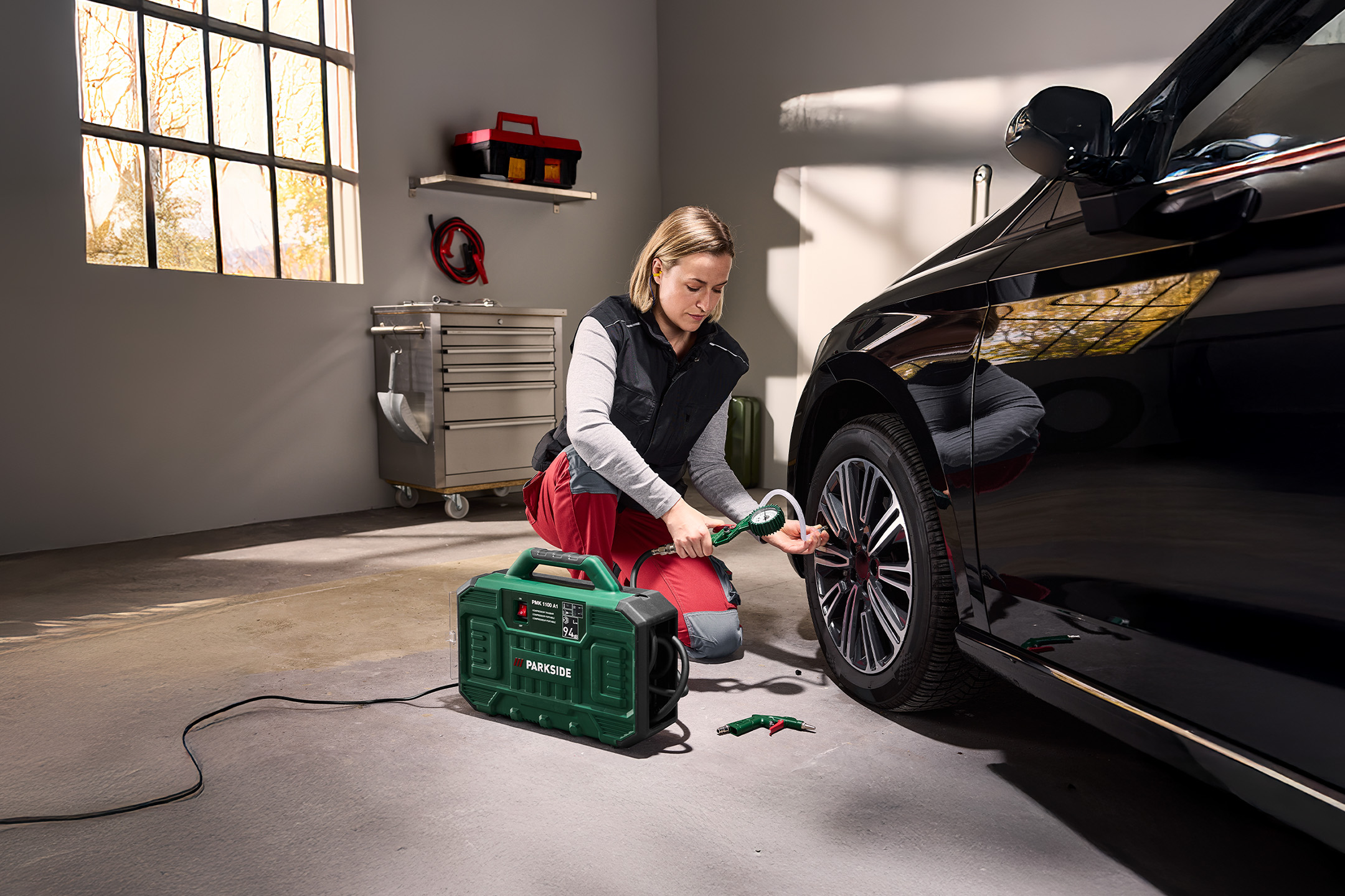Kobieta pompuje oponę samochodu czarnym kompresorem Parkside w garażu.