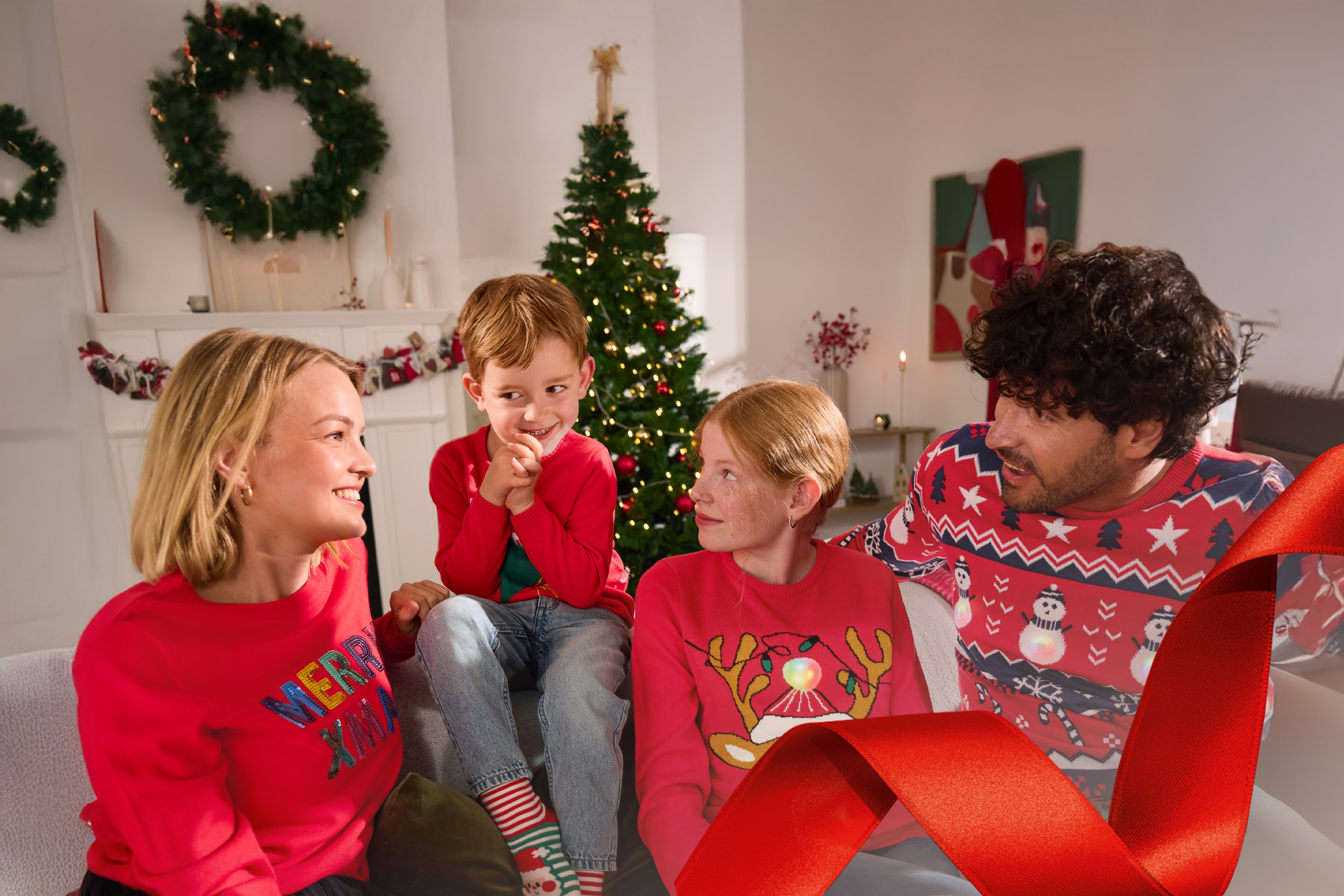 Rodzina w świątecznych swetrach siedzi na kanapie, w tle choinka i dekoracje.