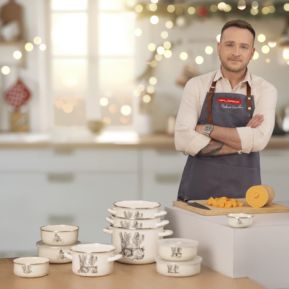 Mężczyzna w fartuchu Florina, naczynia emaliowane z motywami zwierzęcymi, dynia i por na blacie.