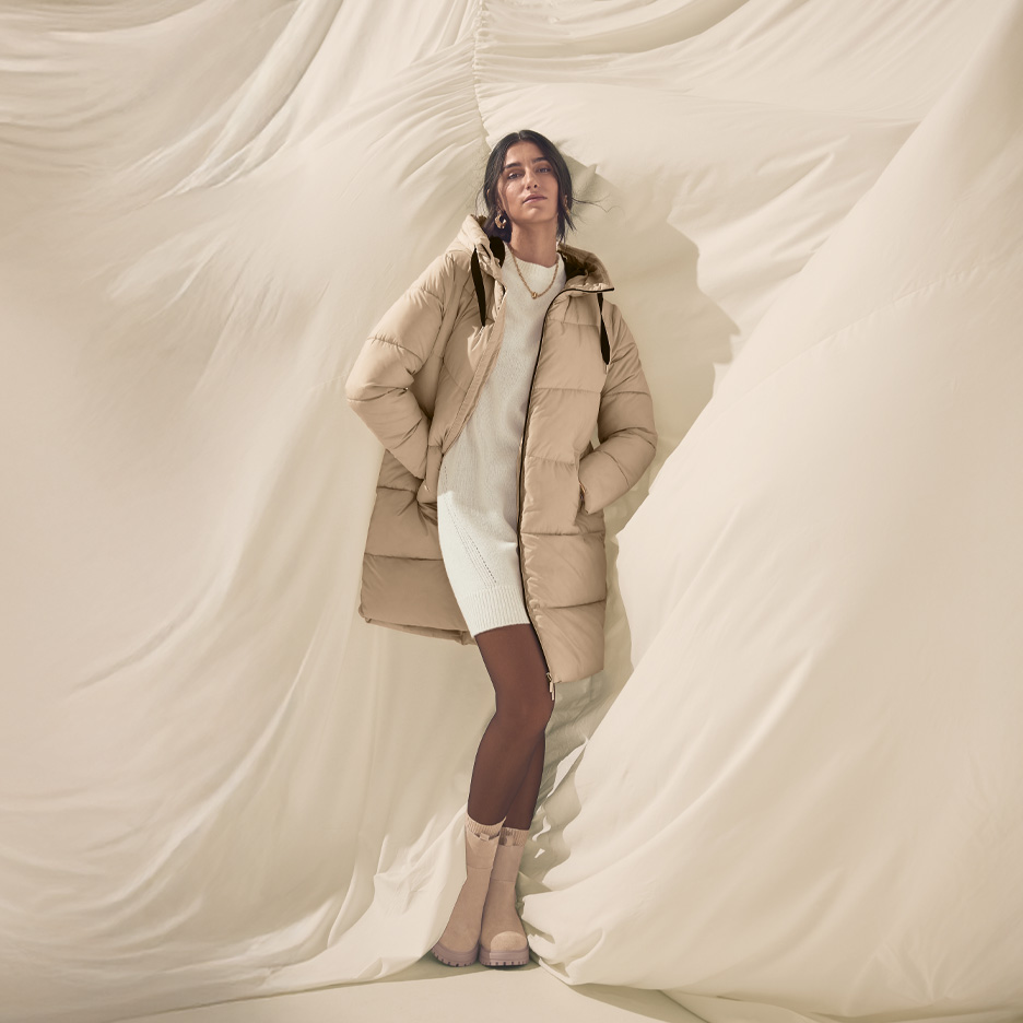 Kobieta w beżowej kurtce puchowej, białej sukience i beżowych butach na tle jasnego materiału.