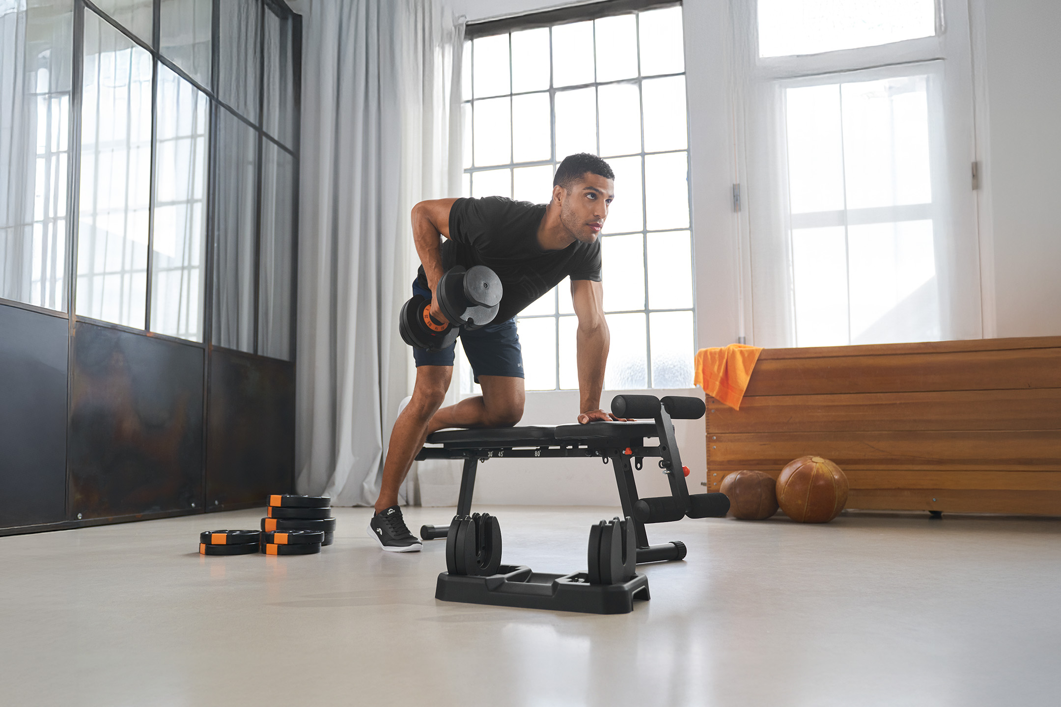 Mężczyzna ćwiczy z hantlami i ławką do ćwiczeń w jasnym pomieszczeniu.
