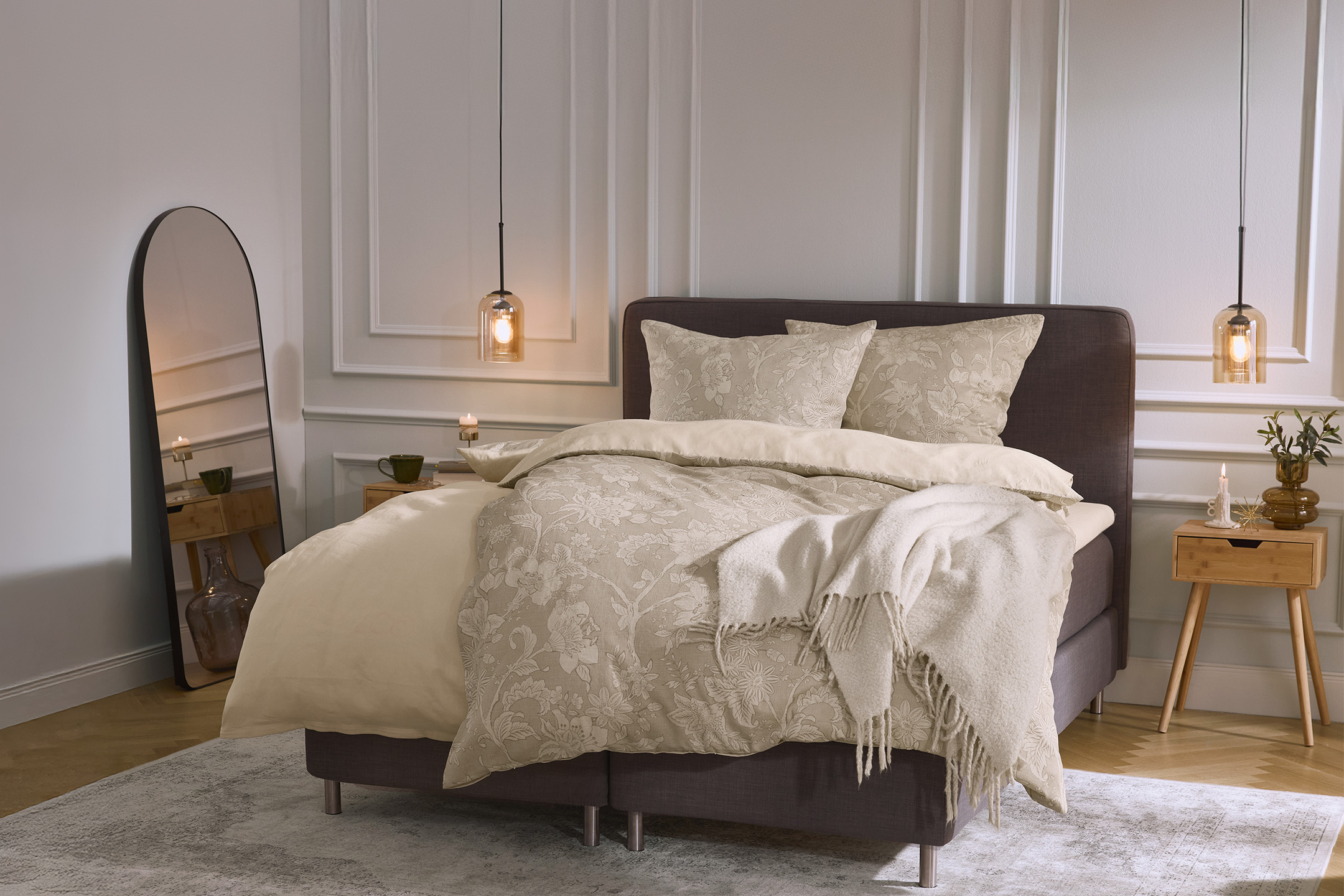 Sypialnia z beżową pościelą w kwiaty, szarym łóżkiem, lustrem i drewnianymi szafkami nocnymi.