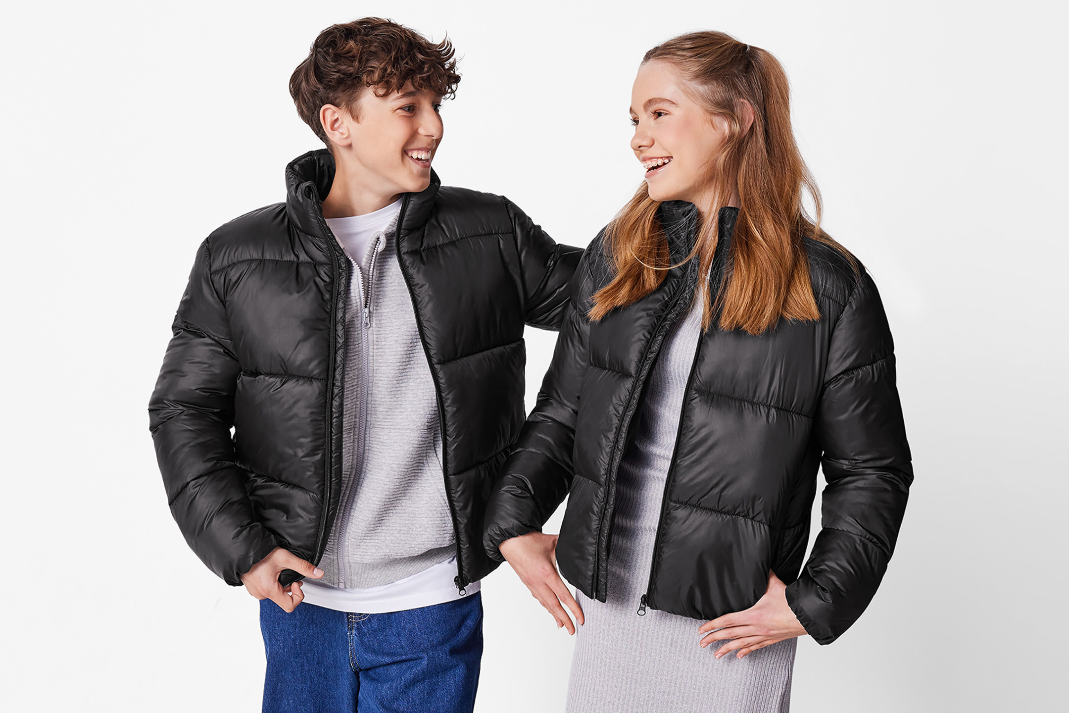 Dwoje nastolatków w czarnych kurtkach puchowych i swetrach, uśmiechających się do siebie.