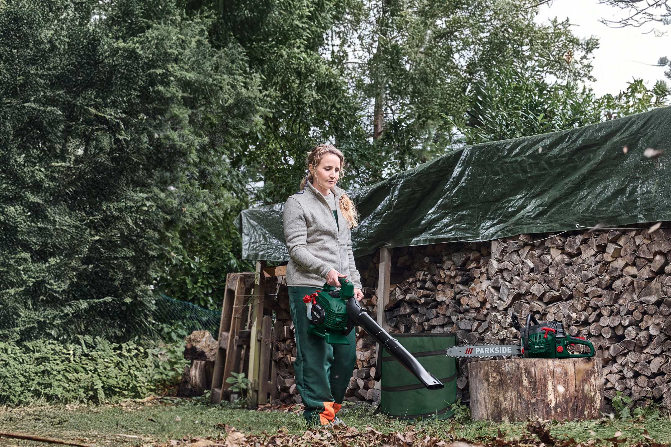 Kobieta z dmuchawą do liści Parkside, piła łańcuchowa Parkside na pniu drewna