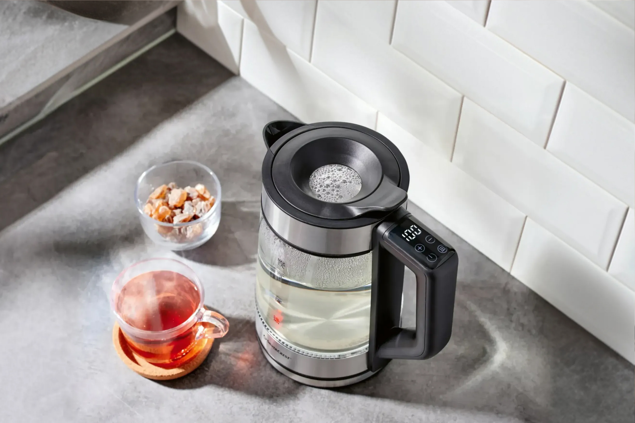 Szklany czajnik Silvercrest z wyświetlaczem, filiżanka herbaty i cukier kandyzowany na blacie.