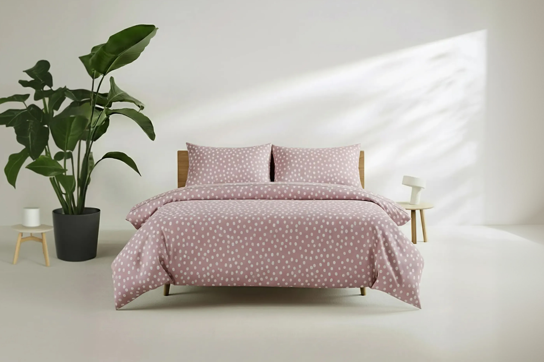 Różowa pościel w białe kropki na łóżku z drewnianym zagłówkiem, obok duża roślina.