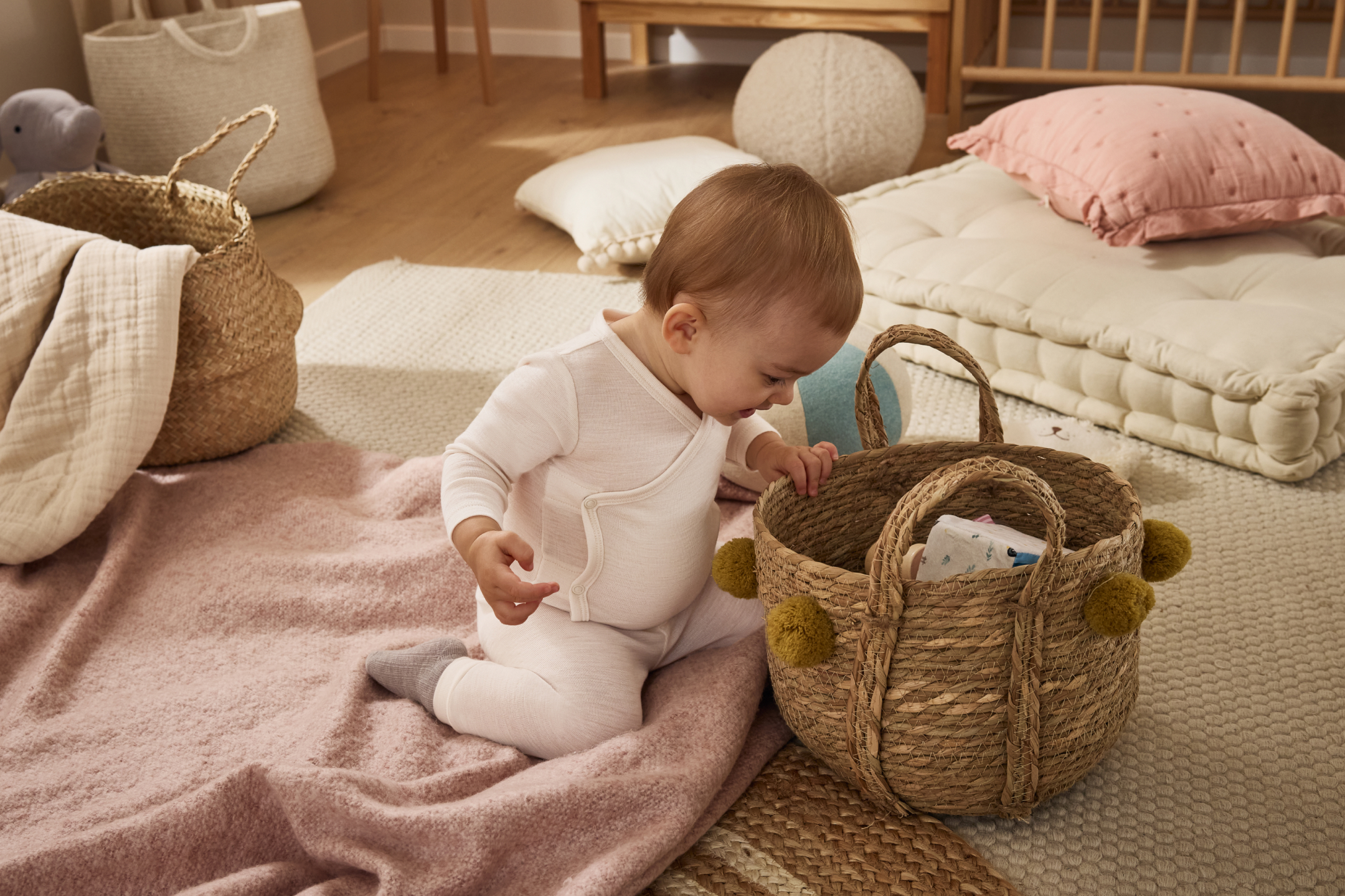 Dziecko w białym body i spodniach, siedzące na różowym kocu obok wiklinowego kosza z pomponami.