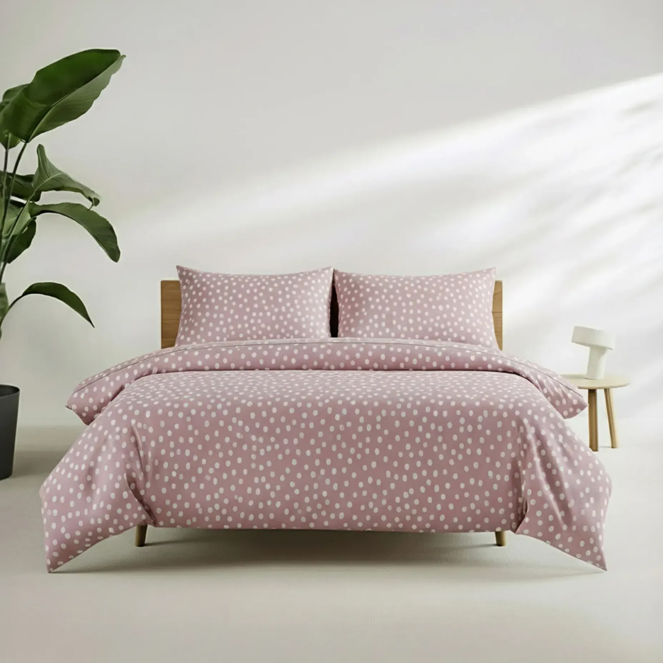 Różowa pościel w białe kropki na drewnianym łóżku, obok roślina i stolik nocny.