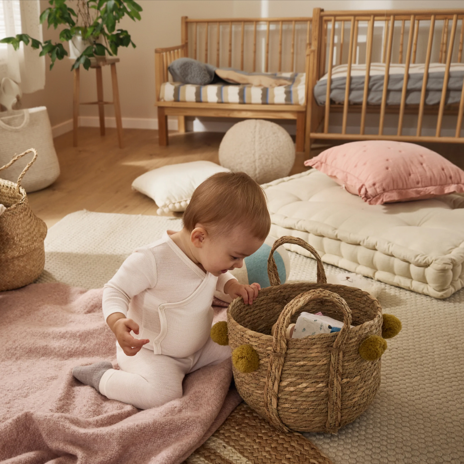 Dziecko w białym ubranku bawi się wiklinowym koszem w pokoju z drewnianymi łóżeczkami.