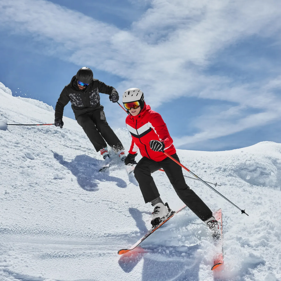 Dwie osoby w kurtkach narciarskich i kaskach zjeżdżające na nartach po zaśnieżonym stoku.