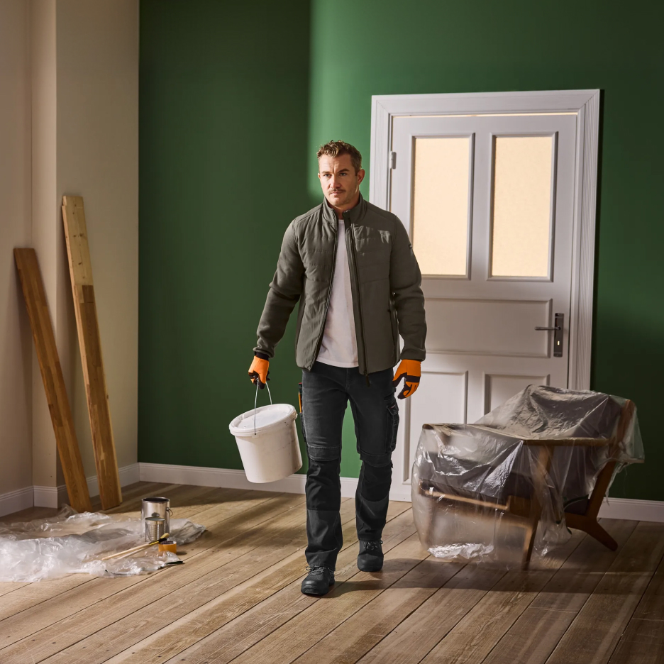 Mężczyzna w kurtce i spodniach roboczych, niosący wiadro z farbą w pokoju do remontu.