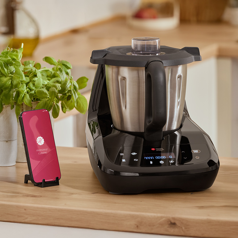 Robot kuchenny Monsieur Cuisine Smart z telefonem z aplikacją na drewnianym blacie.