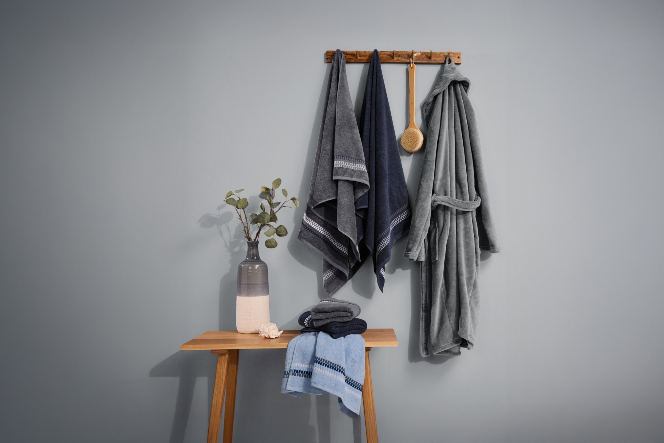 Szare i granatowe ręczniki oraz szlafrok na wieszaku, obok stolik z wazonem i ręcznikami.