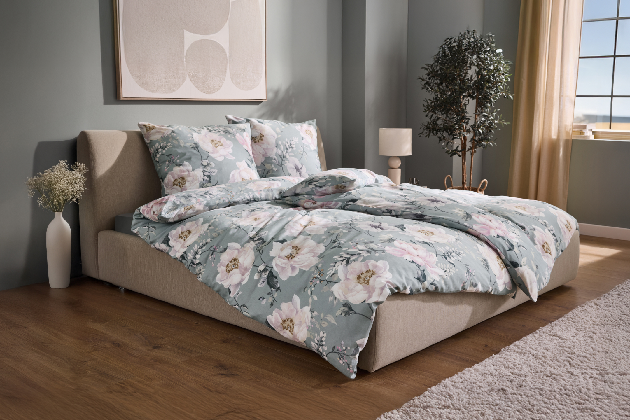 Łóżko z beżową ramą i pościelą w kwiaty, obok wazon z kwiatami i lampka nocna.