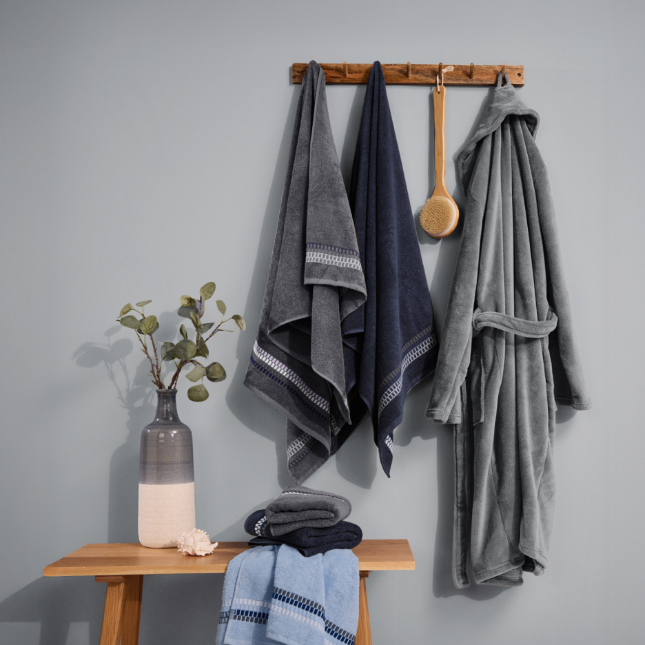 Szare i granatowe ręczniki oraz szlafrok na wieszaku, z wazonem i ręcznikami na ławce.