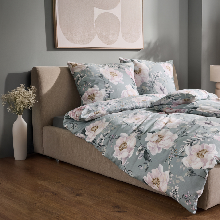 Łóżko z beżową ramą i pościelą w kwiaty, z obrazem na ścianie i wazonem z kwiatami.