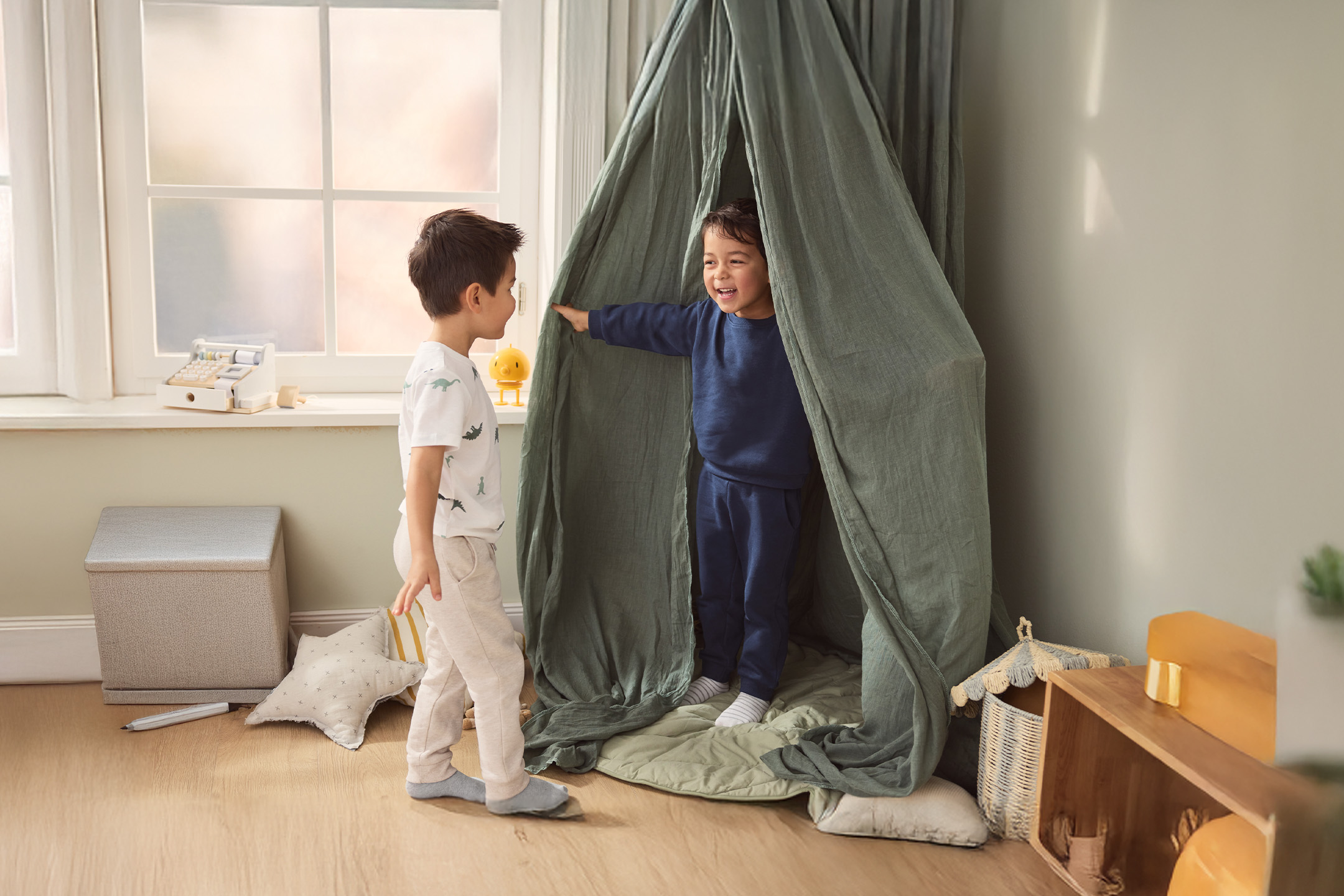 Dwóch chłopców bawi się w pokoju, jeden w namiocie, drugi obok, z drewnianą kasą fiskalną na parapecie.