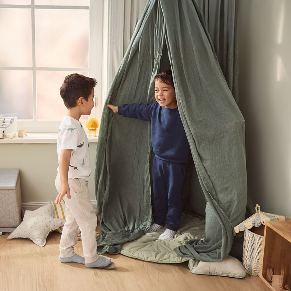 Dwoje dzieci bawi się w namiocie tipi, jedno w piżamie z dinozaurami, drugie w granatowym dresie.