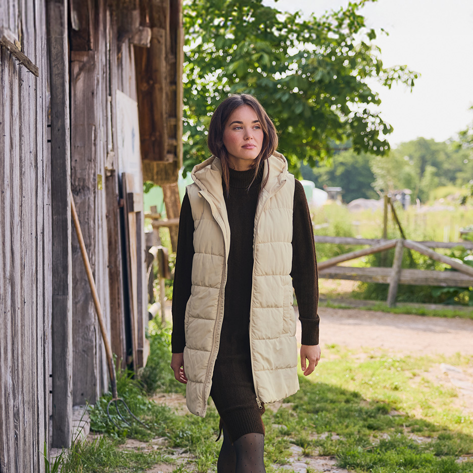 Kobieta w beżowej kamizelce puchowej i brązowej sukience idąca obok drewnianej stodoły.