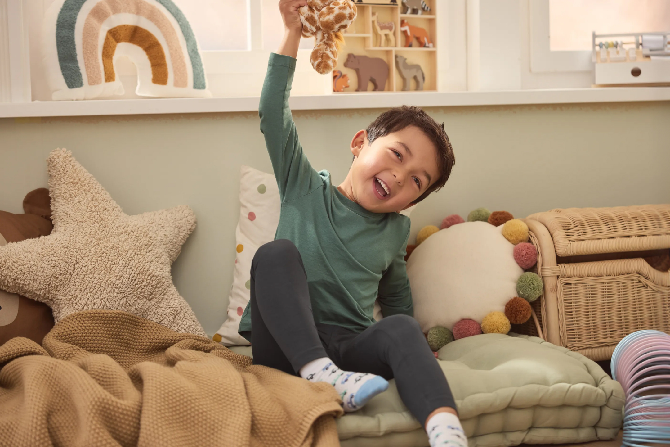 Uśmiechnięty chłopiec bawi się pluszakiem w pokoju dziecięcym z poduszkami i kocem.