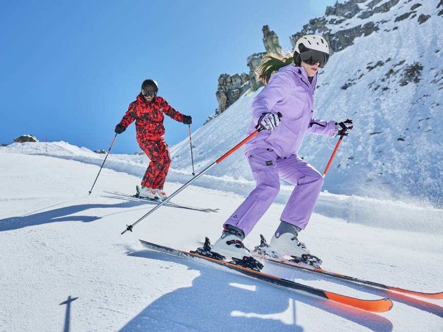 Dwie osoby w kombinezonach narciarskich, kaskach i goglach zjeżdżające na nartach po ośnieżonym stoku.