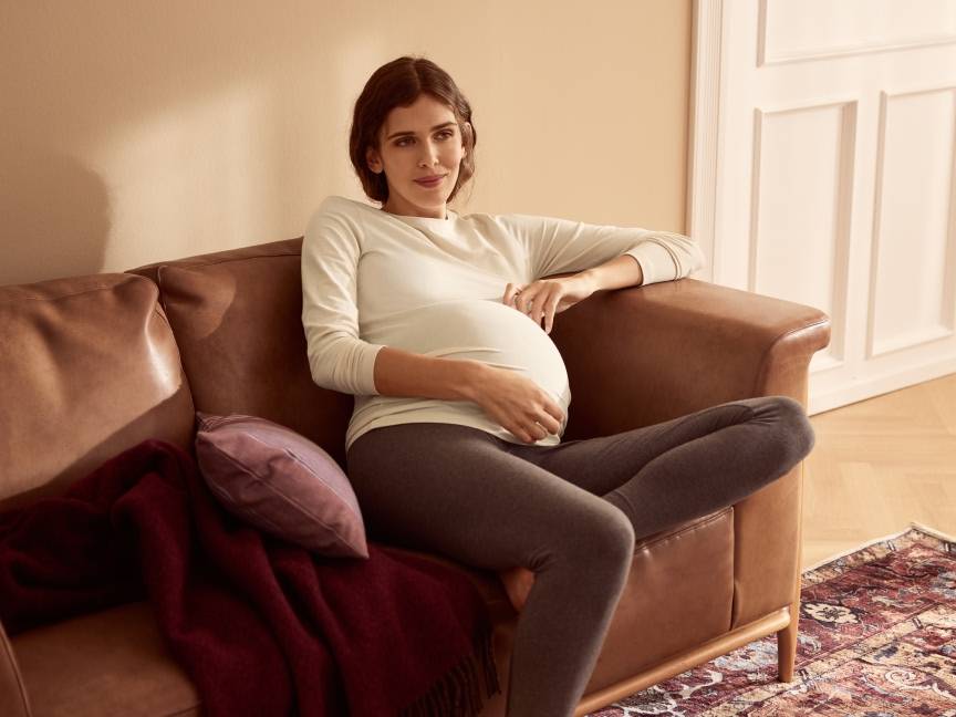Ciężarna kobieta w beżowej bluzce i szarych legginsach siedzi na brązowej kanapie.