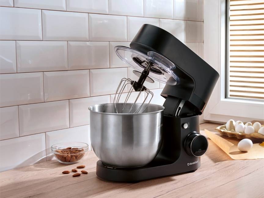 Czarny robot kuchenny z misą ze stali nierdzewnej na drewnianym blacie, obok migdały i jajka.