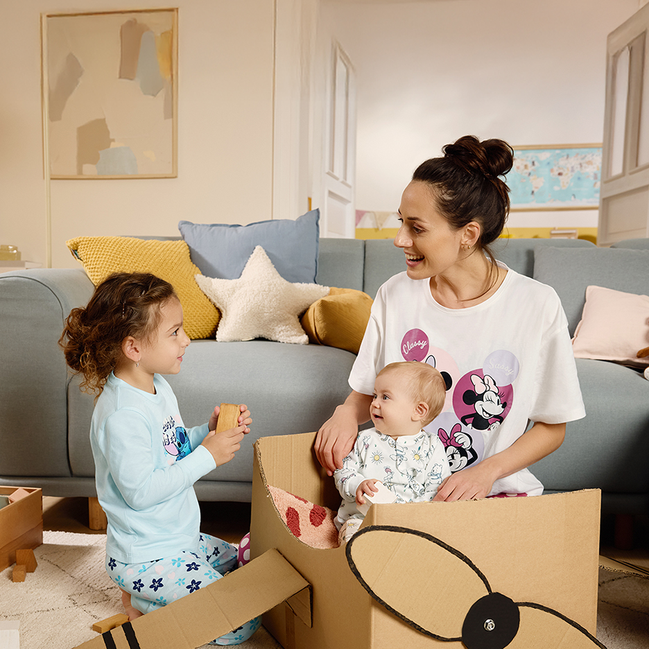 Mama i dzieci w piżamach bawią się w salonie, dziecko siedzi w kartonowym samolocie.