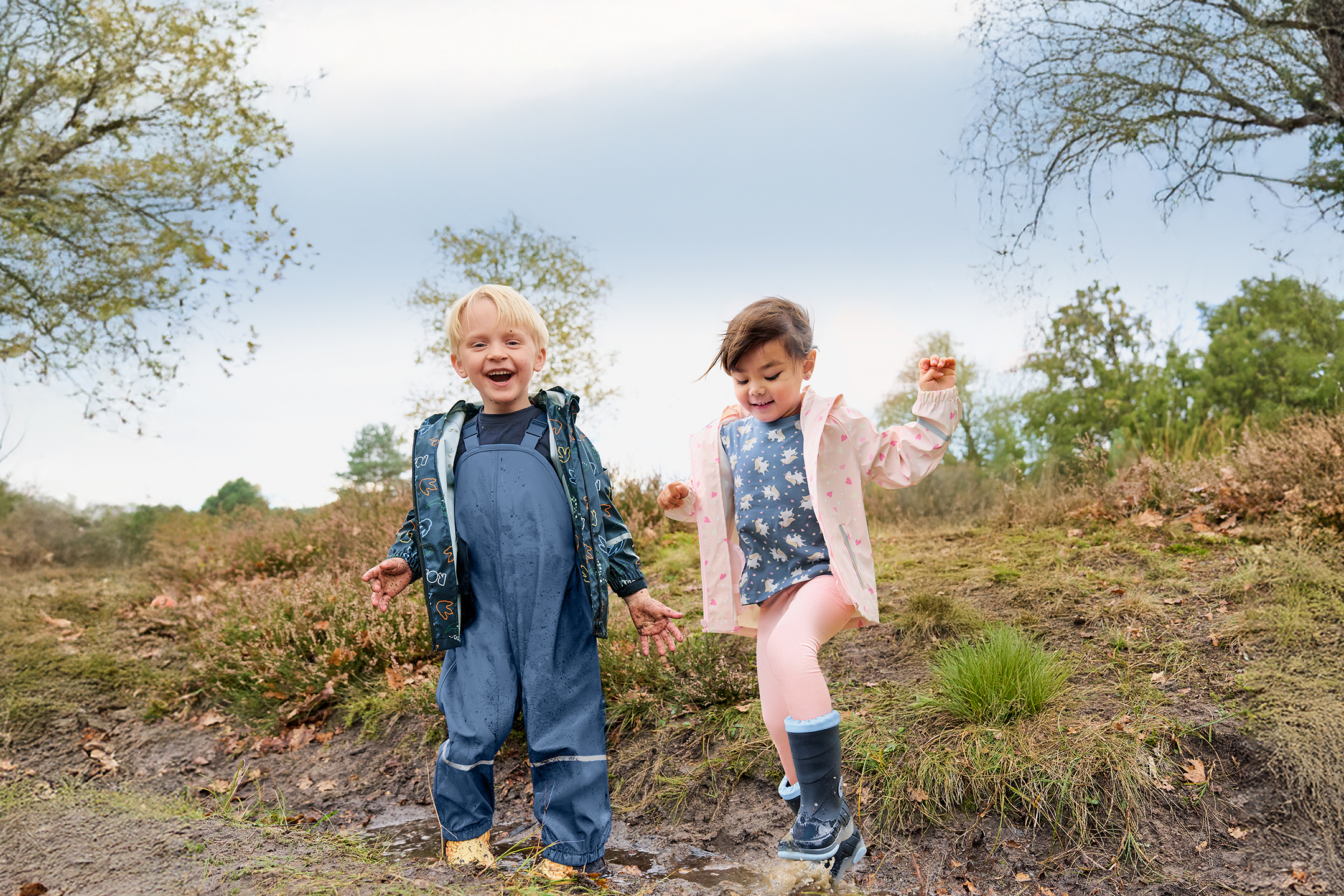 Dzieci w kombinezonach przeciwdeszczowych i kaloszach bawiące się w błocie.