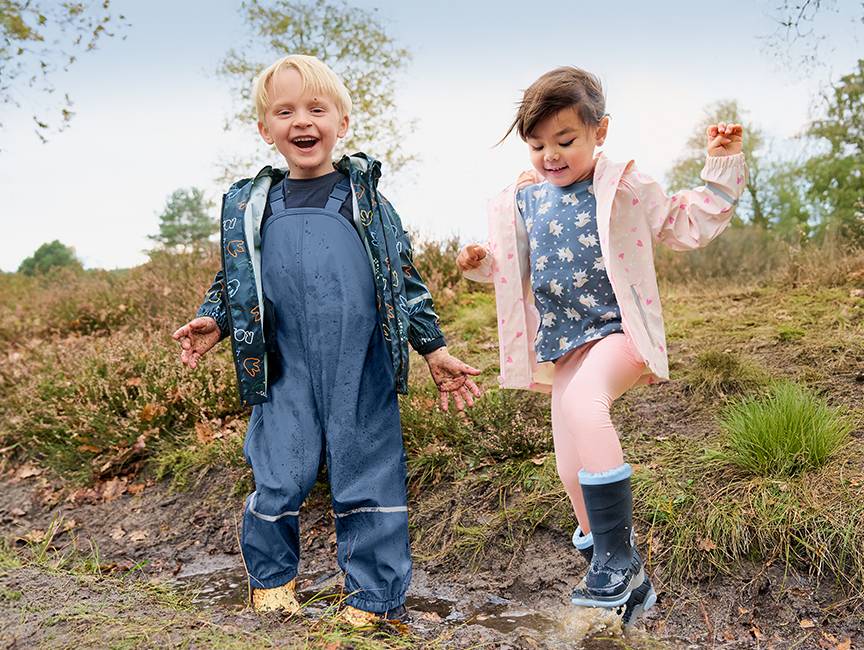 Dzieci w kurtkach przeciwdeszczowych i spodniach na szelkach bawiące się w błocie.